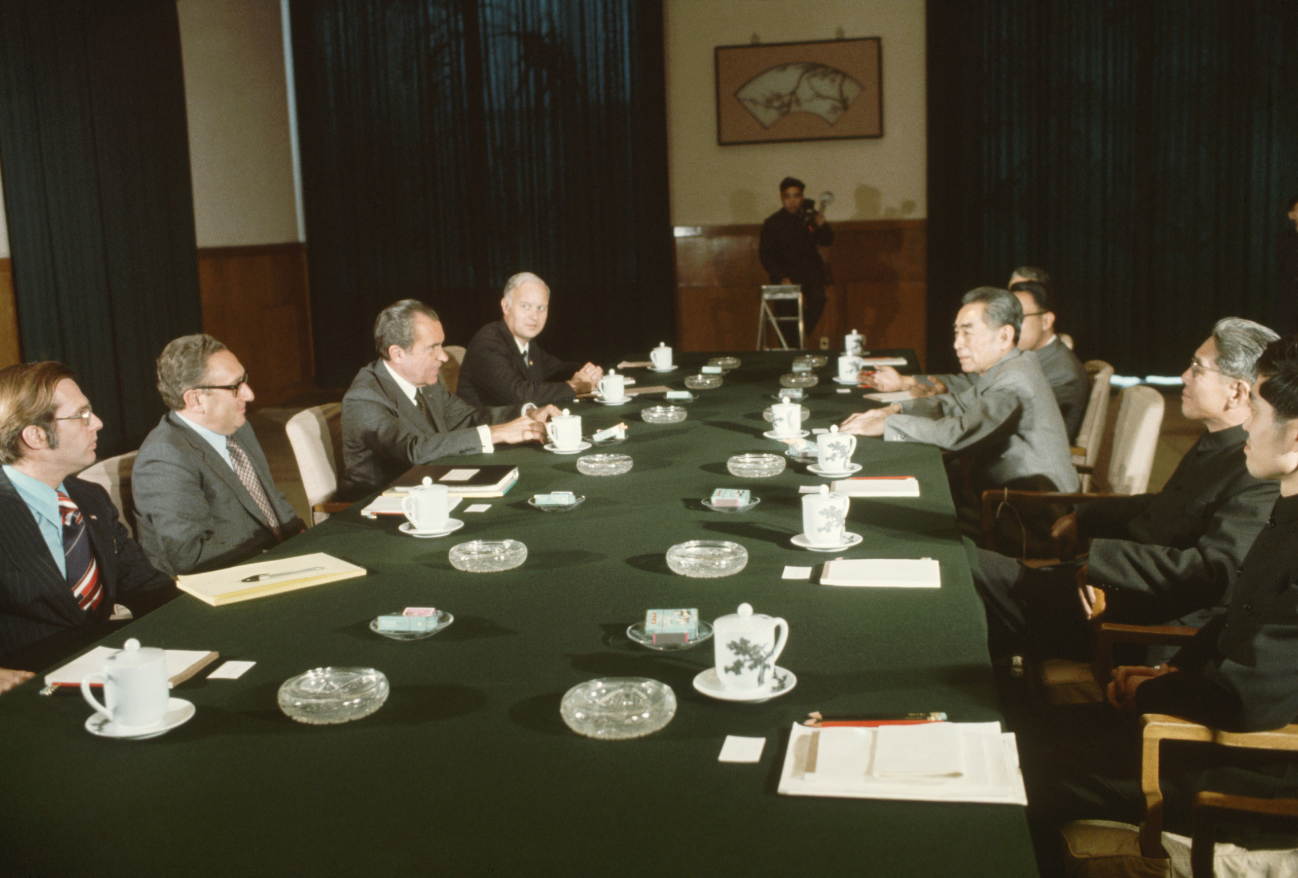 American and Chinese political leaders sit on opposite sides of a table at a meeting during President Richard Nixon's trip to China. On the right are Premier Zhou Enlai (with arms resting on table) and Chinese political leaders. On the other side are American political leaders Winston Lord, Henry Kissinger, Nixon, and William Rogers.